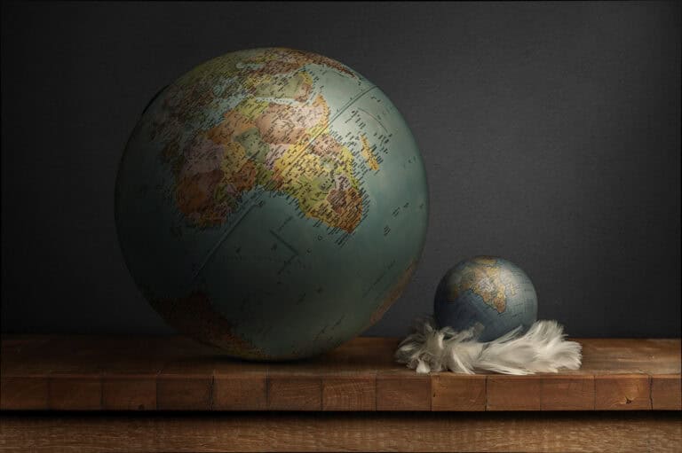 Bodegón conceptual con dos globos terráqueos de distinto tamaño sobre tabla de madera, el pequeño apoyado en plumas blancas, sobre fondo oscuro