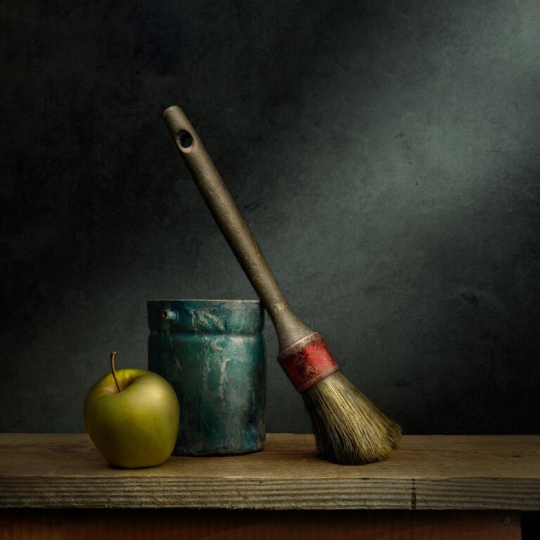 Bodegón fotográfico con brocha vieja, bote de pintura oxidado y manzana verde sobre fondo oscuro
