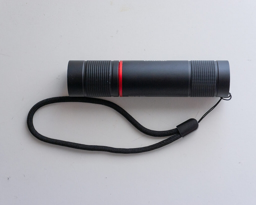 Una linterna táctica de color negro con un detalle de anillo rojo y correa de sujeción, mostrada sobre una superficie blanca.